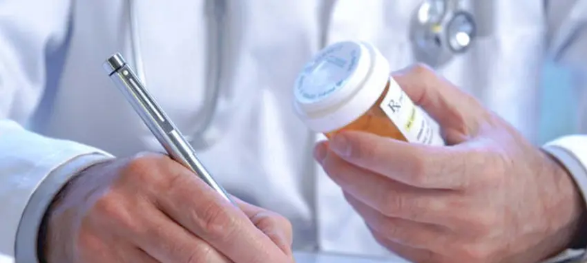A doctor writes a prescription while holding a medicine container during a patient consultation.