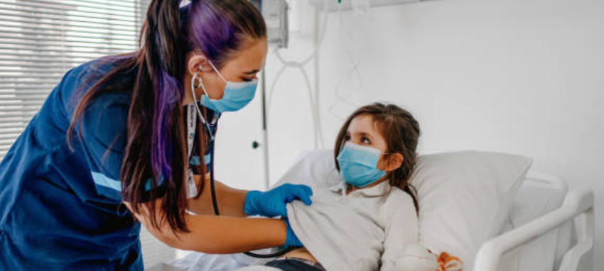 A nurse, wearing a mask and gloves, assists a young girl in a hospital bed, ensuring her well-being