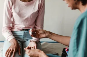 A woman sits on a bed, a man holds her hand and a glucometer to check diabetics.
