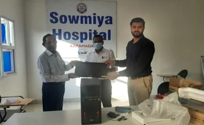 A man giving a PC to another man stands in front of a banner with the words Sowmya Hospital.